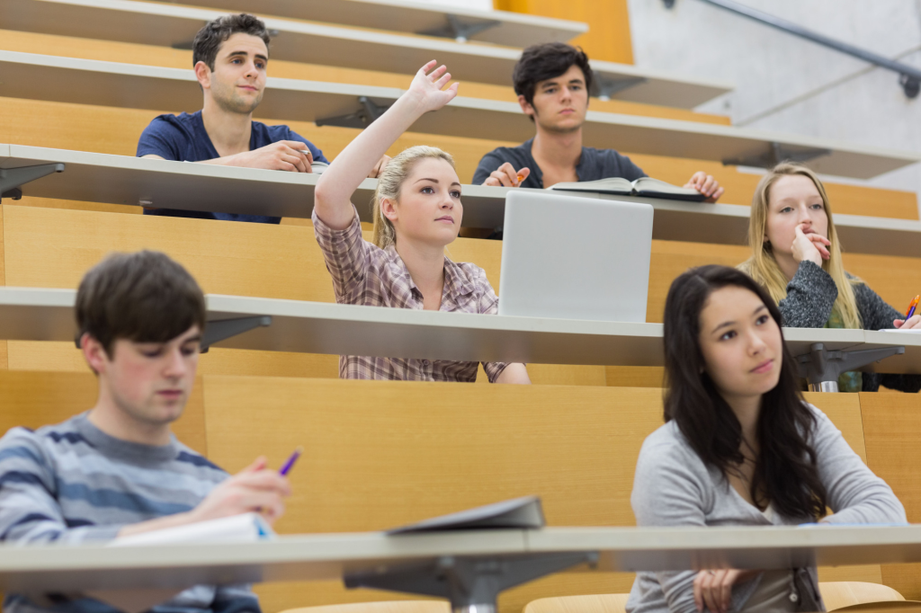 Sinh viên ngồi trong giảng đường đại học, một nữ sinh giơ tay phát biểu trong khi những người khác đang lắng nghe, ghi chép hoặc suy nghĩ.