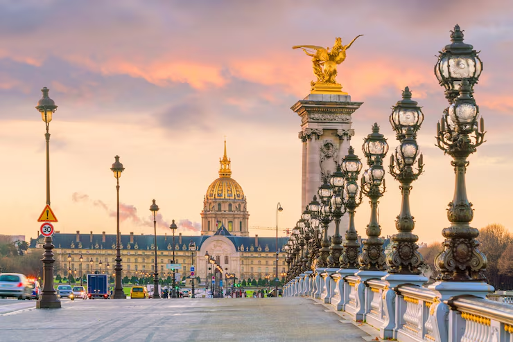Khung cảnh cầu Alexandre III tại Paris, Pháp với dãy đèn cổ điển, tượng vàng và tòa nhà mái vòm phía xa dưới ánh hoàng hôn rực rỡ